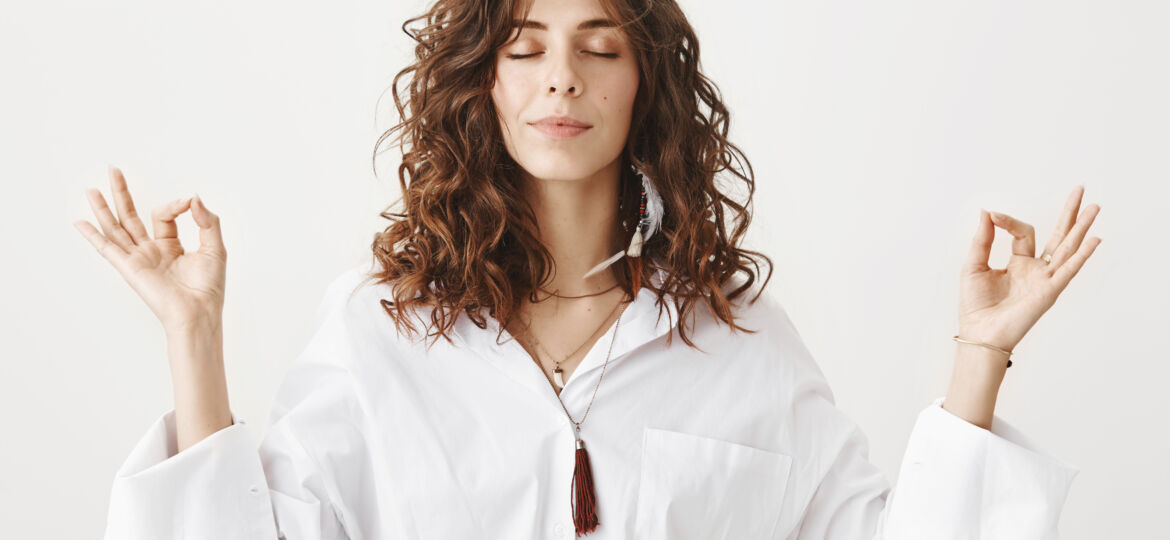 Indoor portrait of calm beautiful caucasian woman meditating in white blouse, smiling while raising hands with zen gestures, standing against gray background. Girlfriend dragged girl to yoga classes